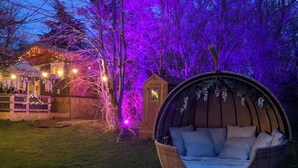 A garden with a wicker swing chair, a wooden shed, and a house with string lights.