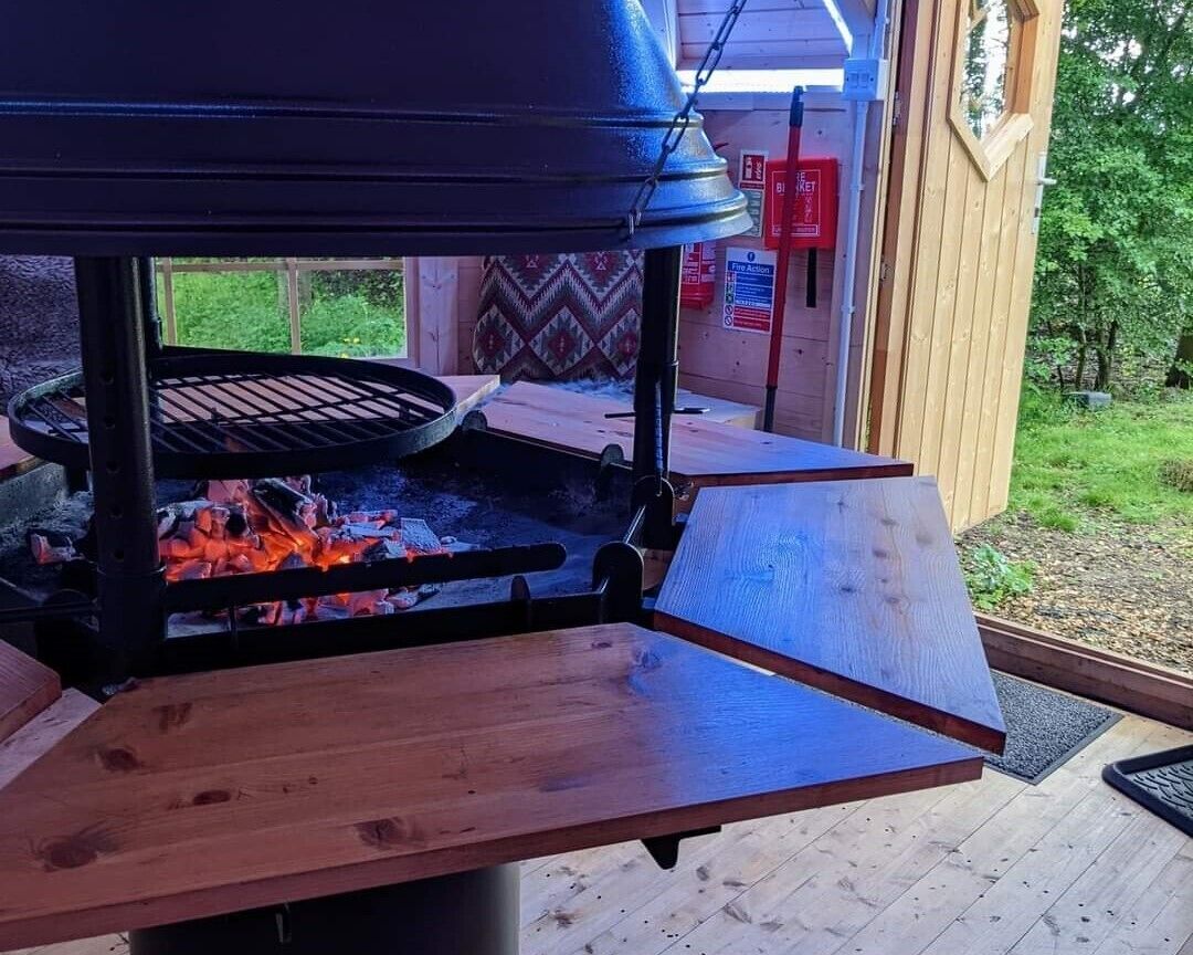 An outdoor grill area with a wooden table and a metal grill, surrounded by a wooden deck and a garden.