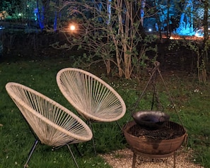 Deux chaises en osier et un récipient en métal posé sur un support, probablement utilisé pour la cuisine, dans un espace extérieur enherbé, de nuit.
