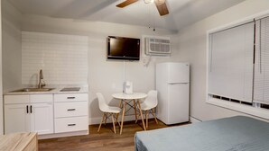 Une cuisine compacte avec des meubles blancs, un évier, une petite table à manger avec des chaises, un ventilateur de plafond, un téléviseur et un réfrigérateur.