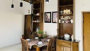Un coin repas avec une table en bois dressée pour quatre personnes, entourée de chaises. On y trouve des lampes suspendues, des étagères en bois où sont disposés divers objets, et une porte sur la droite.