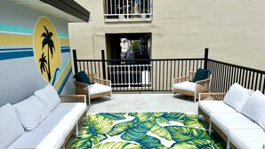A sunlit outdoor seating area with wicker furniture and a tropical leaf-patterned floor. - One Bedroom Suite 107 (Wildwood)