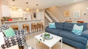 A modern living room with a sofa, coffee table, and kitchen area with bar stools. - Sojourn The Ocean Breeze in Virginia Beach (Virginia Beach)