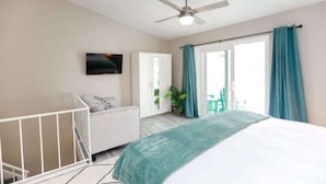 A bedroom with a bed, a white crib, a ceiling fan, a TV, and a sliding glass door with curtains. - Sojourn The Ocean Breeze in Virginia Beach (Virginia Beach)