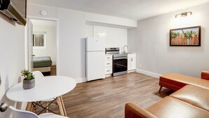 Eine kleine Küche mit weißen Schränken, einem Kühlschrank und einem Herd. Es gibt einen Essbereich mit einem weißen Tisch und Stühlen, sowie ein braunes Sofa. Ein Gemälde hängt an der Wand, und auf dem Tisch steht eine Pflanze.