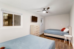 Un dormitorio con dos camas, un ventilador de techo, una ventana, un aparador de madera y un taburete blanco pequeño.