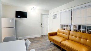 A modern living room with a sofa, a flat-screen TV, a white door, and a window with blinds. - Deluxe Queen One Bedroom Suite 206 (Wildwood)