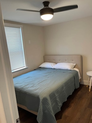 Un dormitorio con una cama, ventilador de techo, ventana con persianas y una pequeña mesa redonda.