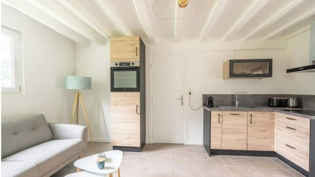 A modern kitchen with wooden cabinets, a built-in oven, and a microwave. There is a white sofa and a small table with a cup on it in the adjacent living area. - Sologne house in the heart of nature, 15 minutes from Chambord Castle! (La Ferté-Saint-Cyr)