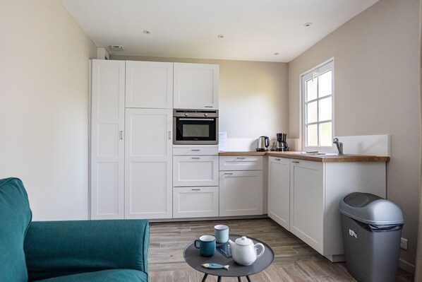 A compact kitchen with white cabinets, a built-in oven, and a coffee maker. There is a small round table with a teapot and cups.