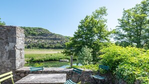 Un espacio de estar al aire libre de estilo rústico con pared de piedra, una mesa y sillas verdes, con vistas a un río y un paisaje montañoso.