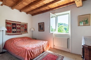 Quarto com teto de madeira, cama com colcha estampada, janela com vista para vegetação, radiador e quadro na parede.