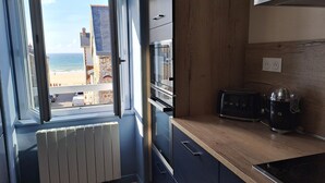 A modern kitchen with a view of the beach and cityscape through an open window.