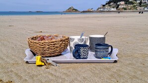A tray with a basket, keys, and patterned mugs on a beach with a view of houses and water in the background. - La Manival - Appartement à 50 m de la plage (Pléneuf-Val-André)