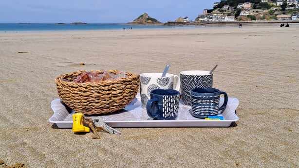 A tray with a basket, keys, and patterned mugs on a beach with a view of houses and water in the background.