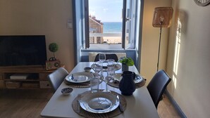 A dining area with a table set for four, a view of the sea through an open window, a clock on the wall, and a plant on the table.