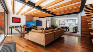 Een ruime woonkamer met een houten plafond, een grote bank, een televisie en een zichtbare eethoek door een glazen deur.