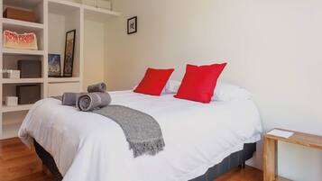 A bedroom with a queen-sized bed, a private bathroom, storage shelves, and a wooden desk.