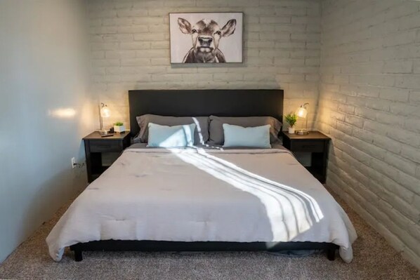 A bedroom with a large bed, two bedside tables with lamps, a brick wall, and a framed picture above the bed.