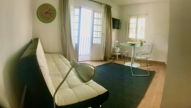 A room with a sofa, a small table with a bowl and a plant, a clock on the wall, and a window with curtains.