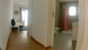 A hallway leading to a bathroom with a toilet, sink, and shower. There is a small table with a vase and a chair in the hallway.
