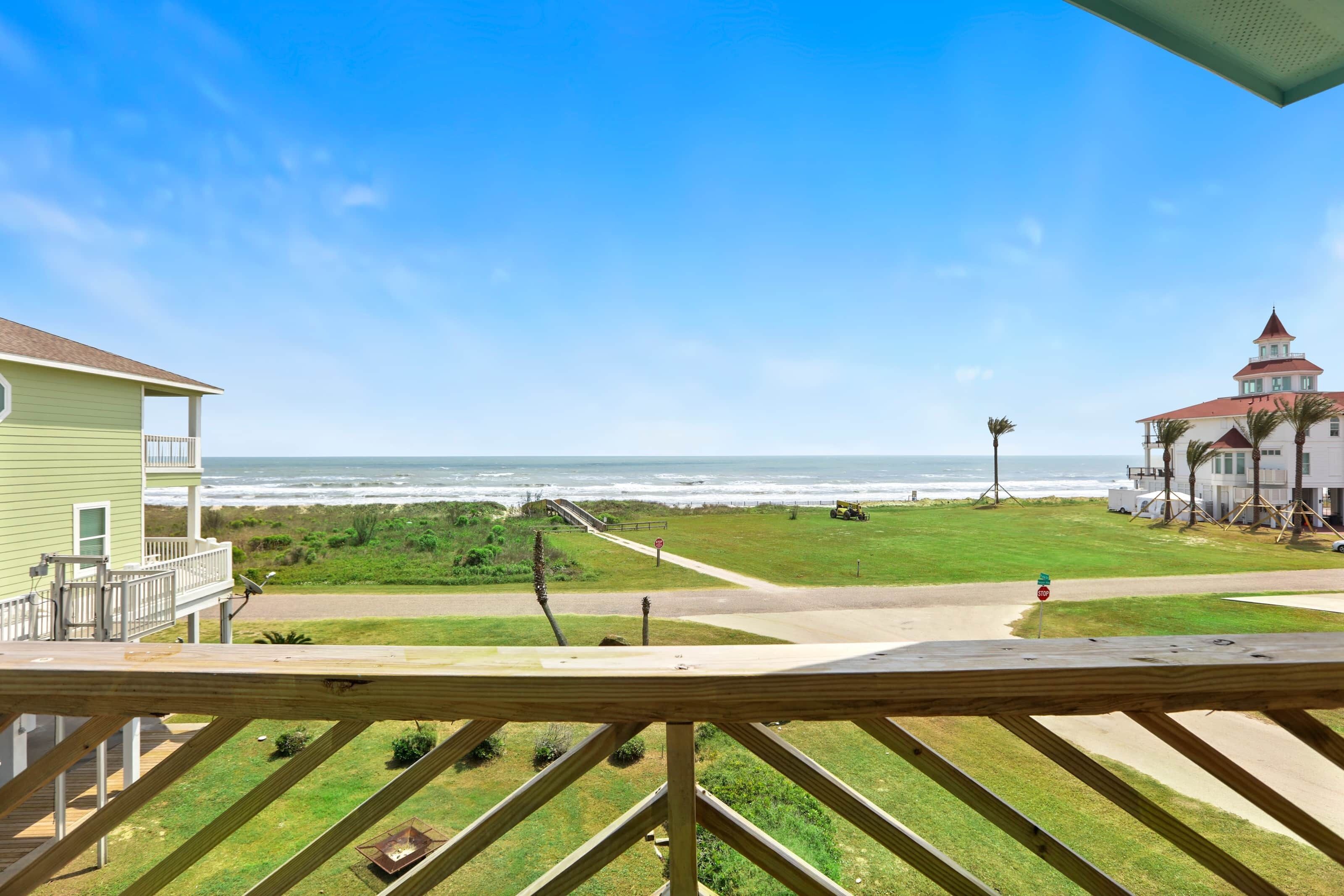 Blick vom Balkon auf einen Strand mit blauem Himmel, einer Rasenfläche und einem Gebäude mit rotem Dach.