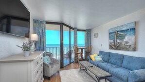 Sala de estar com um sofá azul, uma mesa de centro de madeira, uma poltrona cinza e uma mesa de jantar com cadeiras. Há uma janela grande com cortinas, um quadro de um barco na parede e uma planta em um vaso sobre o balcão.