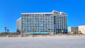 Um hotel grande e com vários andares, de design moderno, situado numa praia sob um céu azul límpido.