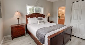 Une chambre à coucher avec un lit en bois, deux tables de chevet avec des lampes, une commode, une fenêtre avec des stores et une porte donnant sur une salle de bain.