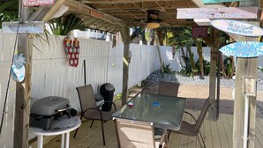Un patio al aire libre con mobiliario de madera, una mesa de vidrio y varios carteles, incluyendo 'Sunset Lounge' y 'Bienvenido'.