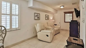 Una sala de estar con un sofá color beige, una silla, un televisor de pantalla plana y un ventanal con persianas.