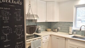 Une cuisine moderne avec des armoires blanches, un panier suspendu, un grille-pain et un tableau noir affichant une liste de cocktails classiques.
