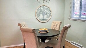 Un coin repas comprenant une table ronde, quatre chaises rembourrées, un lustre et une œuvre d’art représentant un arbre, accrochée au mur.