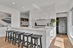A modern kitchen with white cabinetry, a central island with bar stools, a stainless steel refrigerator, and a large window.