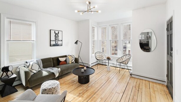 En moderne stue med sofa, sofabord og stol. Stort vindue med persienner, loftslampe og et spejl på væggen.