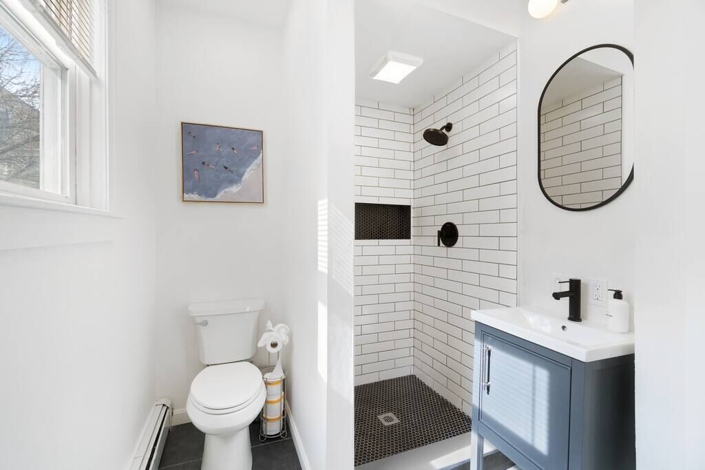 A bathroom with a white toilet, a shower with white and black tiles, a sink with a mirror, and a framed picture on the wall.