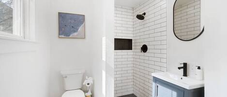 A bathroom with a white toilet, a shower with white and black tiles, a sink with a mirror, and a framed picture on the wall.