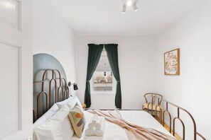 Un dormitorio con cama, ventana con cortinas, una silla e imagen colgada en la pared.