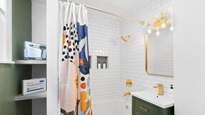 A bathroom with a white bathtub, a shower with a golden showerhead, a green vanity with a white sink, and a mirror with lights. - Perfect for Groups! 3BR★Chic & Modern APT near DT! (Providence)