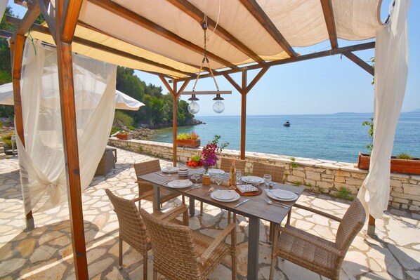 An outdoor dining area with a table set for two, overlooking a body of water. - Nissaki Olive Press Villas-Family House (Nisaki)