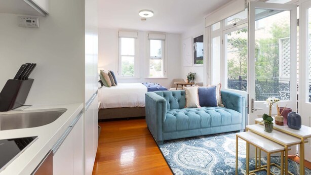 A compact living space with a kitchenette, a bed, a blue sofa, and large windows offering a view of the outdoors. - Intimate Studio Apartment with Balcony in Glebe (Glebe)