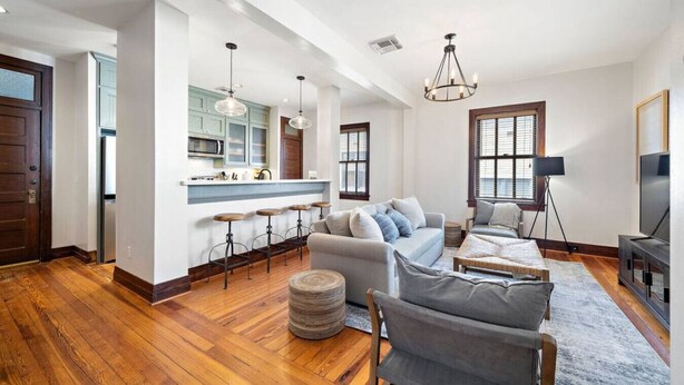 A spacious living room with a grey sofa, a coffee table, and a TV. There is a kitchen area with a bar and stools. - Charming Historic Loft, 3 Blocks from The Strand (Galveston)