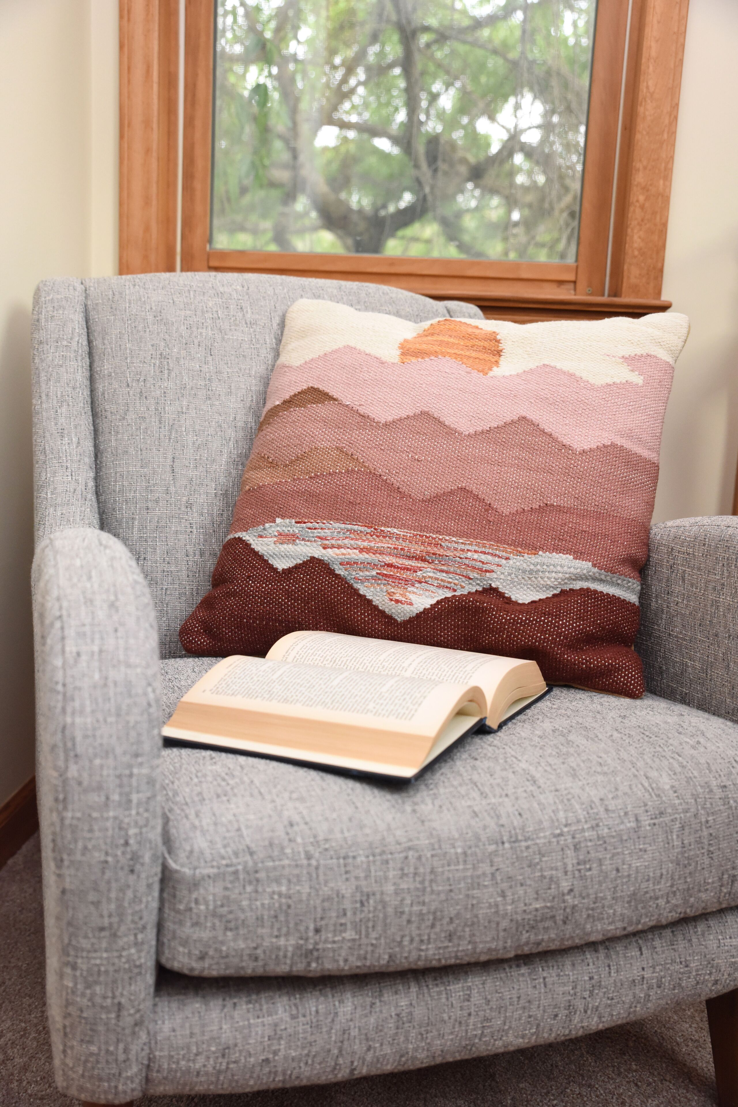 Un sillón gris con un cojín tejido multicolores y un libro abierto encima.