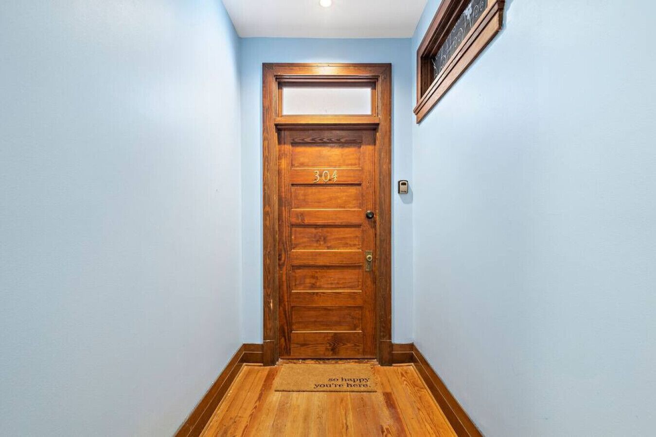 A wooden door with a number plate, a doormat with a welcoming message, and a wall-mounted light fixture.