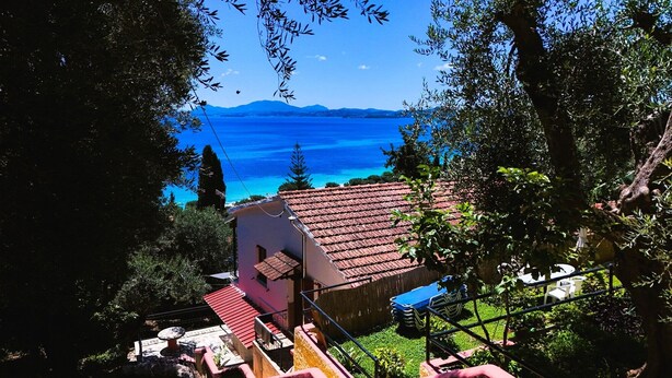 Une maison avec un toit recouvert de tuiles rouges, entourée de verdure, qui offre une vue imprenable sur une mer bleue et des montagnes lointaines.