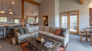 A spacious living area with a large sectional sofa, a coffee table, and a dining table with chairs. There are multiple windows providing natural light and a view of the outdoors. - JHRL - Moose Creek #1, Private Hot Tub (Teton Village)
