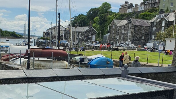A deck with three wooden bar stools and a glass-top table. In the background, there are boats docked at a marina, a street with cars, and buildings. - LOVELY 2 BED APARTMENT PORTHMADOG HARBOURSIDE FANTASTIC VIEWS Ffestiniog trains (porthmadog)