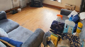A living room with a television, a window with a view of a backyard, a small table with cleaning supplies, and a grey sofa. - LOVELY 2 BED APARTMENT PORTHMADOG HARBOURSIDE FANTASTIC VIEWS Ffestiniog trains (porthmadog)