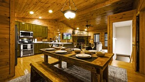 A wooden cabin interior with a dining table set for four, a kitchen area with modern appliances, and a fireplace. - Family Friendly ~ Large Deck ~ Hot Tub ~ Game Room ~ Secluded Getaway (Sevierville)