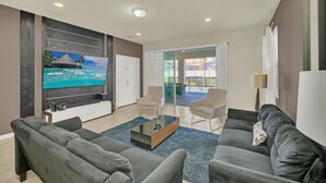 Ein modernes Wohnzimmer mit großem Flachbildfernseher, grauen Sofas, einem Couchtisch und einem Balkon mit Blick auf einen Pool.
